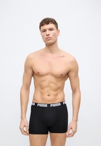 Μυώδης άνδρας που φοράει μαύρα ανδρικά εσώρουχα Puma με λογότυπο στη μέση, στέκεται μπροστά σε απλό λευκό φόντο, κοιτάζοντας μπροστά με ουδέτερη έκφραση.