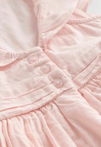 Gros plan sur un vêtement rose pâle avec trois petits boutons translucides et des coutures froncées.