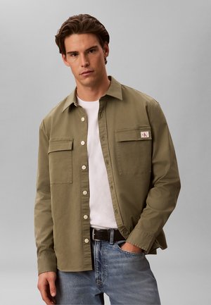 Junger Mann trägt ein olivgrünes Hemd mit Knöpfen über einem weißen T-Shirt und Jeans, steht vor einem einfarbigen grauen Hintergrund.