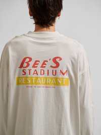 Persona de espaldas lleva una camisa color crema con texto rojo y amarillo que dice "Bee's Stadium Restaurant" y texto más pequeño debajo.