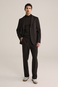 Homme portant un costume marron foncé avec un pull à col roulé et des baskets blanches, debout devant un fond clair uni.