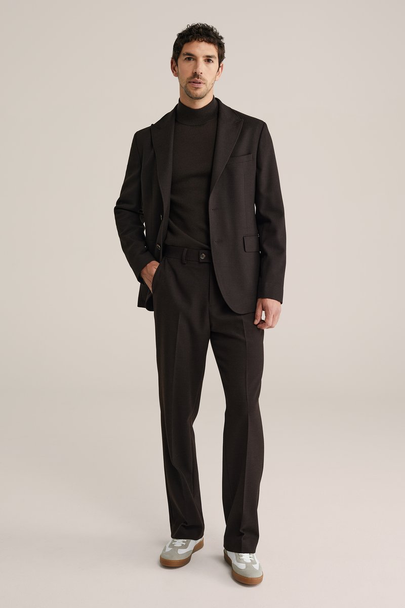 Homme portant un costume marron foncé avec un pull à col roulé et des baskets blanches, debout devant un fond clair uni.