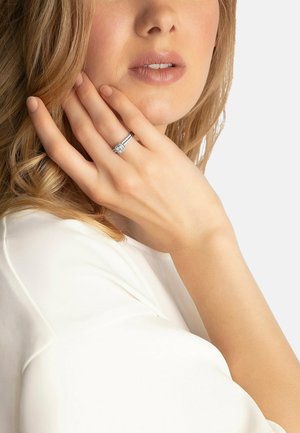 Silberner Ring mit einem zentralen runden Diamanten und zwei kleineren Seitensteinen; getragen an einer Hand mit heller Hautfarbe, vor einem weißen Hintergrund.