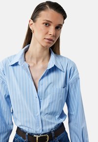 Camicia azzurra con righe verticali bianche, colletto a punta e taschino. Indossata con una cintura marrone e jeans di denim.
