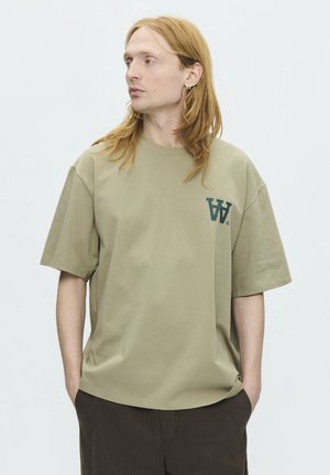 Ung person med langt rødt hår står med hænderne i lommerne og bærer en løs beige t-shirt med et mørkegrønt "W" logo på brystet.