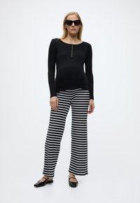 Γυναίκα που φοράει μαύρο μακρυμάνικο τοπ, μαύρο-άσπρο ριγέ παντελόνι, μαύρα φλατ παπούτσια και γυαλιά ηλίου, στέκεται μπροστά σε ένα απλό λευκό φόντο.