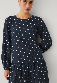 Vestido azul marino con mangas largas, adornado con lunares blancos, escote redondeado y cintura elástica con falda fruncida. Tejido ligero.
