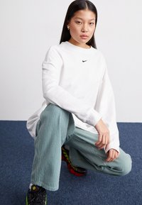 Fehér, hosszú ujjú Nike póló fekete logóval, türkiz és fehér csíkos nadrággal párosítva. A modellen fekete és piros sportcipő van. Kék háttér.