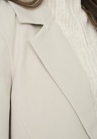 Blazer beige claro con solapa estructurada y textura suave, superpuesto a un suéter de punto acanalado blanco. Detalle en primer plano.