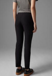 Kvinna i svarta slim-fit byxor, mönstrad kortärmad topp instoppad, och svarta plattformssneakers mot en grå bakgrund.