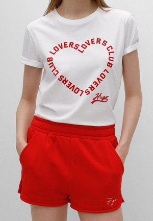 Frau trägt ein weißes T-Shirt mit rotem herzförmigem "LOVERS CLUB"-Schriftzug und rote Shorts, die Hände in den Taschen.