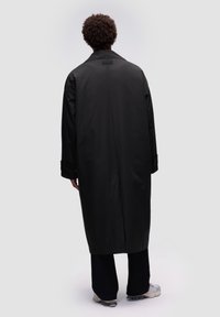 Impermeable negro largo hecho de material suave y resistente al agua. Presenta un cuello, puños abotonados y cae por debajo de la rodilla. Vista trasera mostrada.