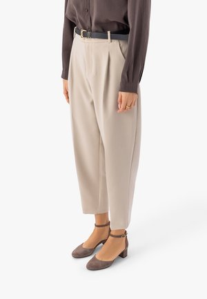 Sieviete, valkājot bēša krāsas augsta jostasvietas bikses ar krokām, melnu jostu, brūnu garā piedurkņu blūzi un brūnas potīšu apavus ar sprādzi un blokpapēdi.