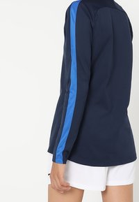 Osoba ubrana w granatową koszulę z długim rękawem z niebieskim paskiem na ramieniu oraz białe spodenki, stojąca na tle jednolitego tła.