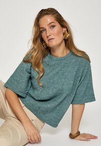 Tyrkysový pletený sveter s krátkymi, širokými rukávmi a okrúhlym výstrihom. Hladká textúra, nosený s krémovými nohavicami a dreveným náramkom.