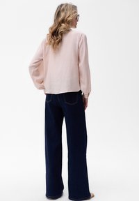 Jasnoróżowa bluzka z długim rękawem w luźnym kroju, zestawiona z ciemnoniebieskimi, szerokimi spodniami dżinsowymi. Bluzka ma delikatną fakturę i boczne rozcięcia.