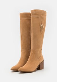 Tan suede knee-high boots with a chunky heel, featuring a side zipper and a strap accent with a small gold-tone buckle. Smooth texture.