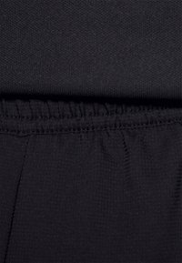 Černá látka s texturovaným povrchem, vybavená elastickým pasem a na švu shromážděnými detaily, které zdůrazňují těsné přizpůsobení.