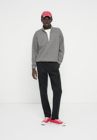 Sweat-shirt gris à fermeture éclair avec un col montant, assorti à un pantalon cargo noir et des baskets rouges. La casquette est rose avec un effet usé.