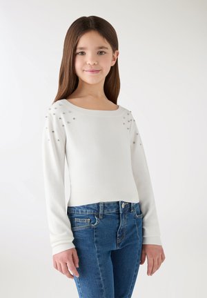 Ragazza con capelli lunghi e castani che indossa un maglione bianco a maniche lunghe con piccole perline sulle spalle e jeans blu, in piedi davanti a uno sfondo semplice.