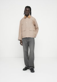 Veste en coton beige avec un col marron contrastant, deux poches avant et des boutons-pression. Associée à un jean gris à jambes larges et des chaussures noires.