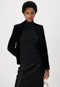 Black velvet blazer with a fitted black turtleneck and glossy black skirt. Model holds a small white handbag. Simple, elegant design.