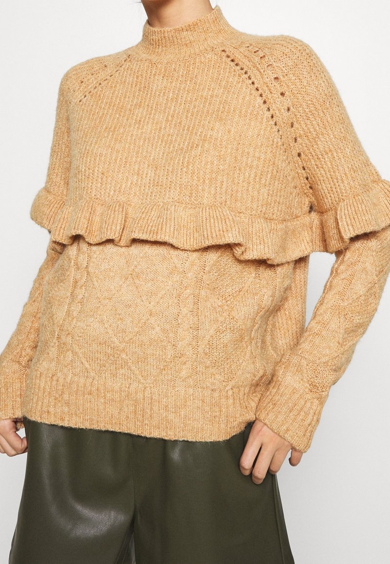 Femme portant un pull beige en maille avec un détail volanté sur la poitrine, associé à un pantalon en cuir vert foncé.