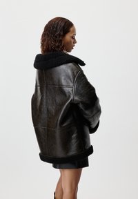 Giacca in shearling di pelle nera con ampio colletto, finitura texturizzata e bordi in shearling a contrasto su polsini e orlo. Vestibilità tailored, lunghezza al ginocchio.