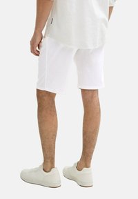 Weiße Leinen-Shorts mit lockerer Passform, versehen mit umgeschlagenen Säumen und Seitentaschen, kombiniert mit weißen Sneakers an einem unbedeckten Bein.