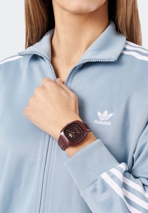 Reloj de pulsera cuadrado morado con esfera negra y detalles dorados. Se lleva puesto en una mano que sostiene una chaqueta azul claro con cremallera y franjas blancas.