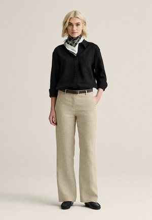 Kvinde med kort blondt hår iført en sort skjorte med knapper, beige bukser med vide ben, sort bælte, mønstret tørklæde og sorte sko, stående.