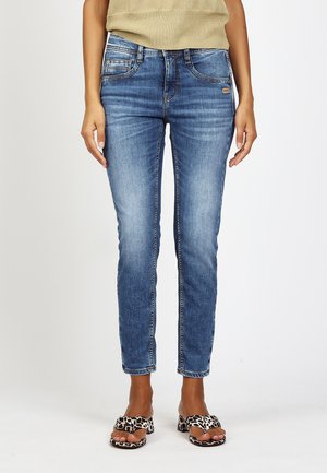 Woman wearing slim-fit blue denim jeans and leopard print open-toe heeled sandals, standing against a plain white background.