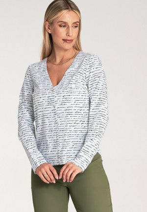 Bluzka z długim rękawem w jasnoniebieskim kolorze, z dekoltem w serek, pokryta czarnym, odręcznym wzorem i detalami w postaci guzików, zestawiona z oliwkowo-zielonymi spodniami.