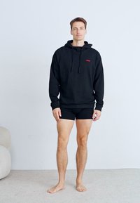 Svart hettegenser med rødt logo, tettsittende svarte shorts, står barbeint på et lyst teppe mot en enkel vegg med minimal bakgrunn.