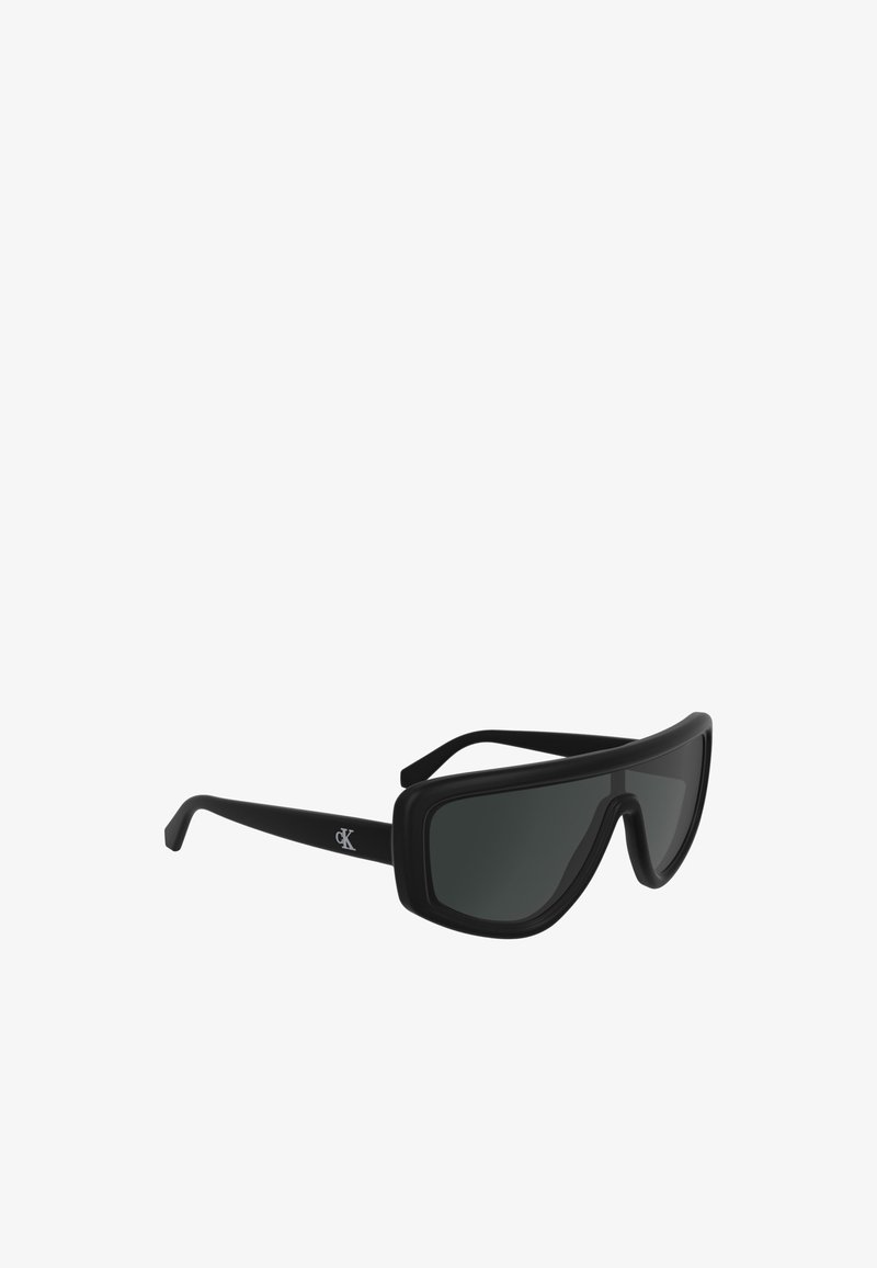 Mustad ümarad mustad prillid, millel on sile ja matte viimistlus. Suured tumedad objektiivid ja eristuv ümberringi disain. CK logo käes.