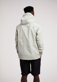 Giacca impermeabile grigio chiaro con cappuccio, caratterizzata da una texture liscia, brandizzazione sottile sul retro e polsini elasticizzati. Abbinata a pantaloni corti neri.