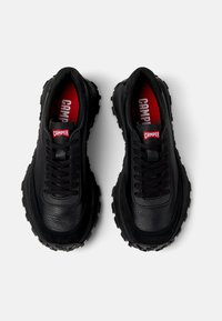 Black sneakers with textured leather and rubber soles, featuring a black lace-up design and red "CAMPPER" logo on the tongue.