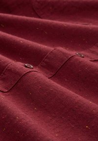 Tissu bordeaux avec un motif en chevron, comportant des éclats de jaune, rouge et bleu. Deux boutons sur le col et des poignets repliés visibles.