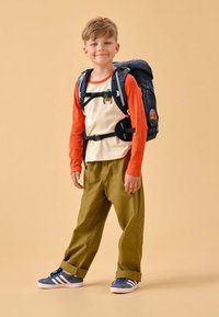 Lo zaino presenta una base nera con un motivo colorato; il bambino indossa una maglietta arancione a maniche lunghe, pantaloni beige e scarpe blu.