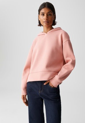 Frau in einem hellrosa Kapuzenpullover und dunkelblauen Jeans, stehend mit einer Hand in der Tasche vor einem schlichten Hintergrund.