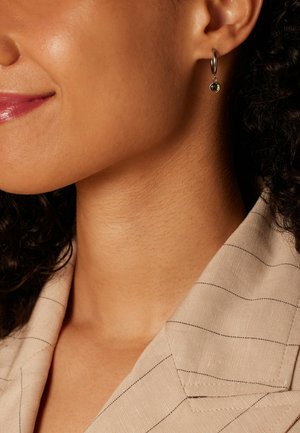 Zilveren oorring met een kleine groene edelsteen hanger, gecombineerd met een beige shirt met streepjes, dat gladde texturen en subtiele details laat zien.