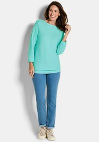 Minzgrüner Pullover mit weitem Ausschnitt und 3/4-Ärmeln, kombiniert mit blauen Jeans und hellbeigen Sneakers. Weicher, leichter Stoff.