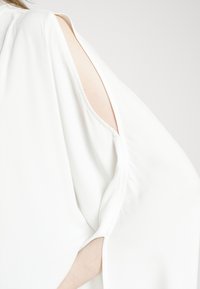 Blusa bianca leggera con maniche aperte, caratterizzata da una texture liscia e una leggera caduta. Le fessure laterali migliorano la traspirabilità e il design.