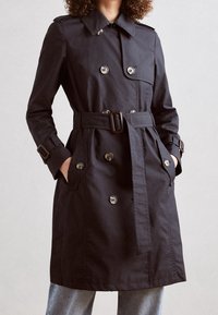 Svart trench coat med dubbelknäppning, justerbara ärmslut och ett matchande bälte. Har sidofickor och en klassisk krage.
