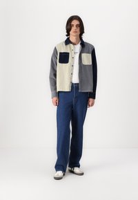 Jachetă din denim cu blocuri de culoare în crem, gri închis și bleumarin, cu două buzunare frontale. Purtată peste un tricou alb și asortată cu blugi albaștri.