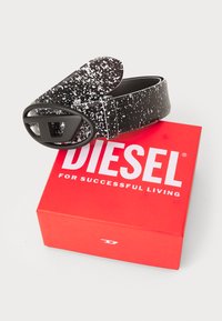 Ceinture à pois noir et blanc avec une boucle noire mate, reposant sur une boîte rouge étiquetée "DIESEL POUR UNE VIE RÉUSSIE."