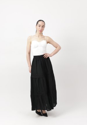 Kobieta stojąca z ręką na biodrze, ubrana w białą, bezrękawnikową bluzkę, długą czarną spódnicę z koronkowymi detalami i czarne szpilki na tle jednolitego białego tła.