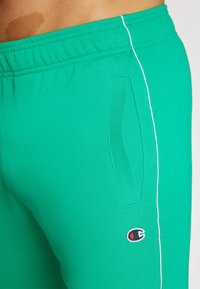 Green cotton sweatpants featuring an elastic waistband, side pockets, and a contrasting white stripe along the side. Logo on the leg.