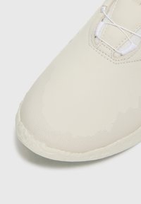 Chaussure de sport beige clair avec une surface lisse et texturée, comportant des motifs en pointillés subtils et des lacets blancs, conçue dans une forme épurée.