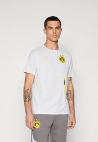 Hvid kortærmet skjorte med gule accenter og en lynlåslomme. Inkluderer Puma-logoen og et cirkulært sort logo med gule detaljer.